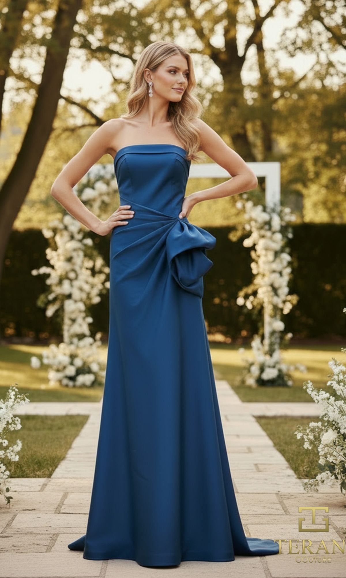 A woman poses outdoors in a strapless teal Long Formal Dress (Terani 2111P4019) with a large side bow, surrounded by white floral arrangements.